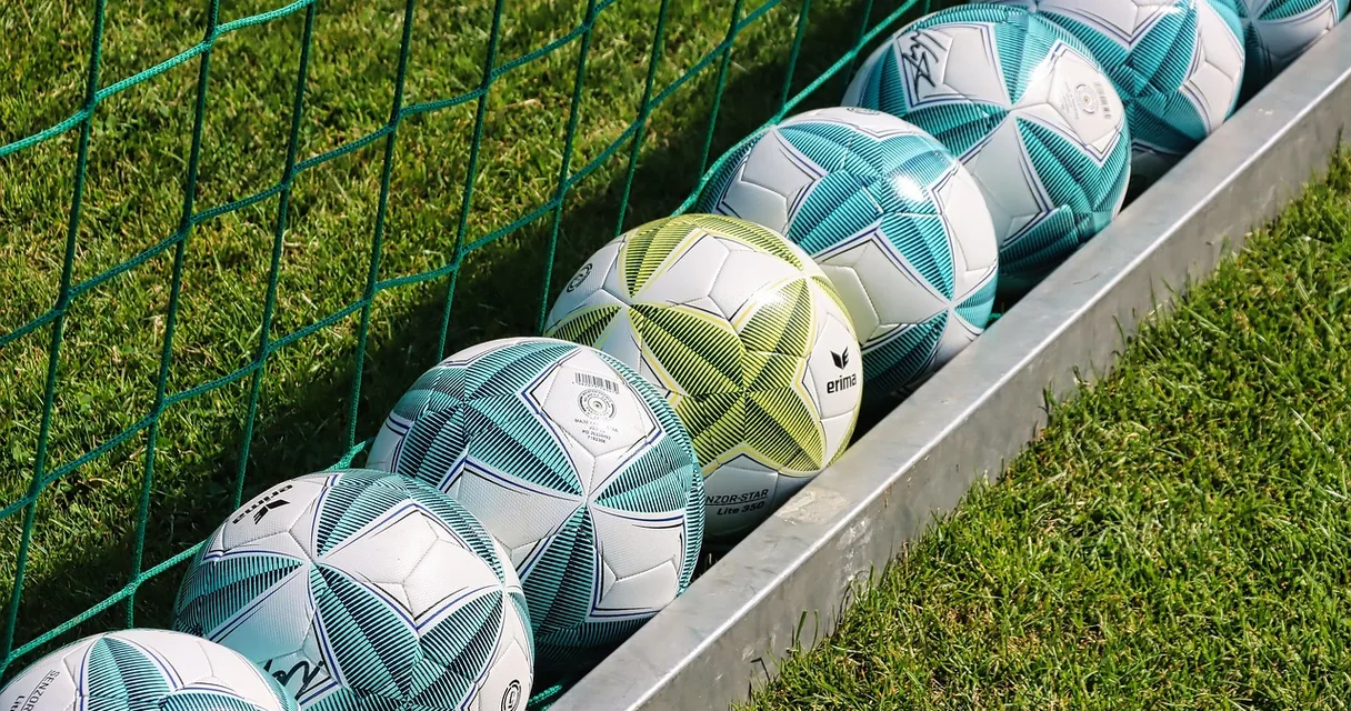 Bolas de futebol enfileiradas ao lado de uma rede em gramado, ilustrando o dedo atingido pela bola